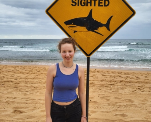 Carolina am Mona Vale Beach neben einem Schild, das vor Haien warnt